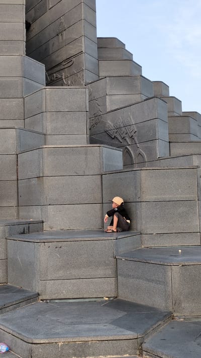 A low-angle shot shows a boy squatting on a large, gray, concrete stepped structure that appears to be a monument in an outdoor setting. The monument is constructed of multiple gray concrete blocks in a stepped pattern. The boy wears a cap, dark clothing, and sandals, and he is looking down, seemingly focusing on something on the ground. Engraved designs are visible on some of the concrete blocks. The background shows a bit of blue sky. The image suggests an urban environment. It is Vietnam's Unknown city.