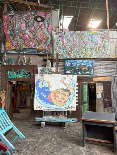 The image depicts an art studio space in Tejakula, Indonesia, likely an open-air or semi-enclosed area given the visible wooden beams and corrugated metal roof. The space is filled with various artworks, primarily paintings and some sculptures.
In the foreground, a light blue, weathered wooden chair is partially visible on the left, angled towards the center. To the right of the chair, a small, dark wooden desk with a slanted top and a lower shelf stands on casters. Its surface is empty.
Dominating the center of the image is a large painting on an easel. The painting features a stylized depiction of a person lying in a hammock, rendered in bold blue and yellow outlines with red and orange accents. The year "2013" is clearly visible in the bottom left corner of this painting, and a signature is present on the bottom right. The easel itself is a rustic, distressed blue wooden structure with wheels.
Behind the central painting, and slightly to the left, is an open doorway framed by intricately carved wooden elements. Above the doorway, a turquoise sculpture of a stylized human figure in a swimming or floating pose is mounted. Inside the doorway, a yellow cabinet is visible, and further back, a painting of a human figure is hanging.
Above the doorway and cabinet, a large, dark-framed painting hangs on the wall. This painting shows a group of figures in dynamic poses, rendered with broad brushstrokes in shades of pink, blue, and brown. To the right of this large painting, another horizontally oriented painting is displayed. This one depicts a rural scene with a small house, palm trees, and two swans in water, all rendered in bright, vivid colors.
Further to the right, more artworks are visible, including smaller sculptures lined up on a shelf above the doorway and what appears to be a framed print or painting of a face. The walls are constructed from a mix of materials, including wood planks, possibly bamboo, and concrete. The floor is paved with irregular, dark-colored stones. The overall impression is that of a working art studio or gallery, filled with eclectic pieces and displaying a bohemian or rustic aesthetic. The lighting suggests it is daytime, with natural light filtering in from above.