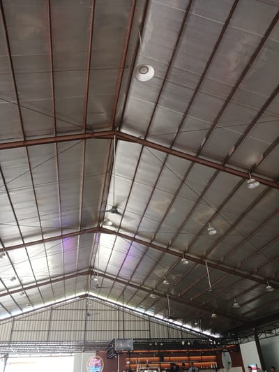 The image captures the interior of a large, open-air establishment with a high, corrugated metal roof supported by exposed metal beams. Several industrial-style pendant lights and ceiling fans are visible, attached to the roof structure. Natural light streams in from the sides, creating bright areas on the ceiling. In the background, a bar area is partially visible with people standing around. A neon sign that reads "SP4 Kopi" is also present, indicating the establishment is likely a cafe or coffee shop. The overall impression is a spacious, casual setting, possibly a restaurant or entertainment venue. The location is specified as Bireuen, Indonesia.