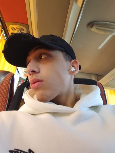 A young man wearing a ripped black baseball cap and white hoodie looks to the left. He has an earbud in his right ear. The setting appears to be inside a bus or a similar vehicle, evidenced by the overhead compartment and the seat to his left. Sunlight streams in, lighting his face.