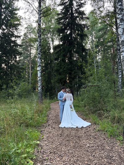A newly married couple stands on a wood chip path in a dense forest in Bakhtiyary, Russia. The bride, wearing a long-sleeved white wedding dress and holding a bouquet, faces her groom. He is dressed in a light blue suit and has his arm around her. The path is surrounded by lush green trees and foliage, with tall birch and pine trees dominating the landscape. The lighting suggests it is late afternoon or early evening, with soft natural light filtering through the trees.