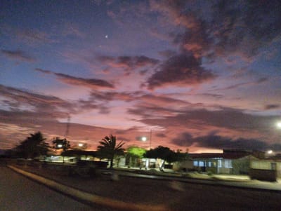 The image captures a dusky sky over a residential area in El Espinal, Venezuela. The sky is a gradient of deep blue and purple, with streaks of pink and orange clouds reflecting the setting sun. A few bright points, possibly stars or distant lights, are visible in the upper part of the sky. In the foreground, a dark, paved road or pathway stretches into the distance. To the left and right of the road are low-lying buildings, appearing to be houses, with lights on inside some of them, suggesting it is dusk or early evening. There are scattered trees and vegetation around the buildings. A utility pole with some wires is visible behind the buildings. The overall scene depicts a quiet, rural or suburban setting at twilight.