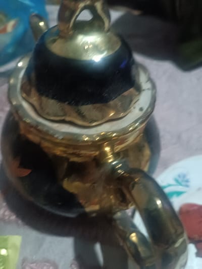 A close-up shows a partially visible, ornate teapot. Its handle is gleaming gold, as is the trim around the lid. The lid's upper portion is a dark, possibly black, material with a reflective sheen. The body of the teapot is a combination of the dark material and gold accents. Some surrounding objects and surfaces are slightly out of focus.