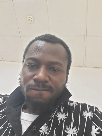 A close-up image depicts one adult male with dark skin, short dark hair, and a dark beard and mustache, whose forehead appears slightly reflective. He is dressed in a black collared shirt featuring white palm trees and vertical white stripes, worn over a visible white t-shirt. The individual is looking directly forward. The background shows a white ceiling constructed of square acoustic tiles, with a circular, off-white smoke detector or fire alarm device centrally positioned. The wall behind the subject is plain white. The image contains no visual information to corroborate the location as Sabon Gari North, Nigeria.