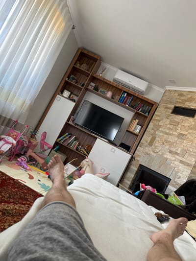 The image shows a view from the perspective of someone lying on a bed, looking towards a living room. The room features a large wooden bookshelf filled with books, decorative items, and framed pictures. A flat-screen television is mounted on the bookshelf. To the right of the bookshelf is a fireplace with a stone facade. In the foreground, a person's legs and feet are visible, suggesting they are in a relaxed position. Scattered on the floor are children's toys, including a doll in a stroller, suggesting the presence of children in the household. A large white curtain covers a window on the left side of the image. The overall environment appears to be a home interior, likely in Băcioi, Moldova.