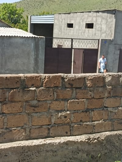 In Nor Geghi, Armenia, a man in a white polo shirt and blue jeans stands to the right of a dark brown gate. His head is turned towards the right, and he is looking down. Partially obscured by the man, another person's head is visible, also looking down. The building behind them is made of concrete with two small, dark rectangular windows. A decorative metal grate is in front of the gate. The foreground is dominated by a brick wall made of rough, reddish-brown bricks. In the background, a green, grassy hill rises under a clear sky.