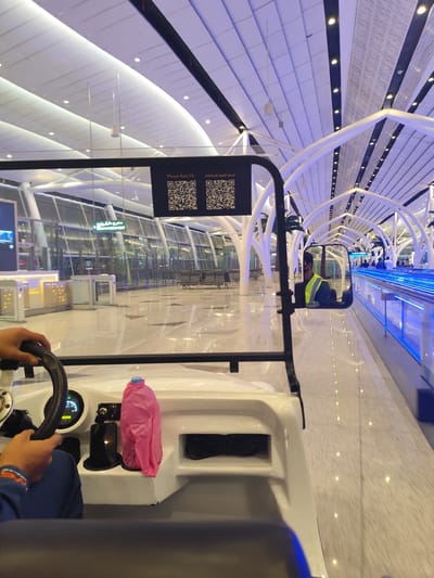 This image captures an interior view from inside a white electric golf cart, likely in a modern airport terminal in Jeddah, Saudi Arabia. The perspective is from a passenger or driver, with hands visible on the steering wheel in the lower left.
The setting is characterized by its spaciousness and contemporary architecture. High, vaulted ceilings feature a combination of white arched structures and panels, illuminated by recessed lights that emit a soft blue-purple glow, particularly noticeable along the ceiling and a long moving walkway on the right. The floor is a light-colored, reflective surface.
In the mid-ground, a black sign attached to the golf cart's frame displays "Please Rate Us" in English and "الرجاء تقييم لخدمتنا" in Arabic, along with two QR codes, inviting feedback. To the left, large glass walls partition the area, and a green "EMERGENCY EXIT" sign (مخرج طوارئ in Arabic) with an arrow is visible. Beyond the glass, benches suggest a waiting area.
A rearview mirror on the golf cart's right side reflects a person wearing a high-visibility yellow safety vest, likely the driver or an attendant, indicating staff presence. Further down the terminal, more white architectural arches, artificial palm trees, and distant people are visible alongside the moving walkway, which is also illuminated with blue light.
On the golf cart's dashboard, a pink plastic bag or container is visible next to a digital display showing "0 KM/H" and other indicators. The bright, clean environment, advanced infrastructure, and specific signage are consistent with a large, modern airport. The overall impression is one of efficiency and comfort.