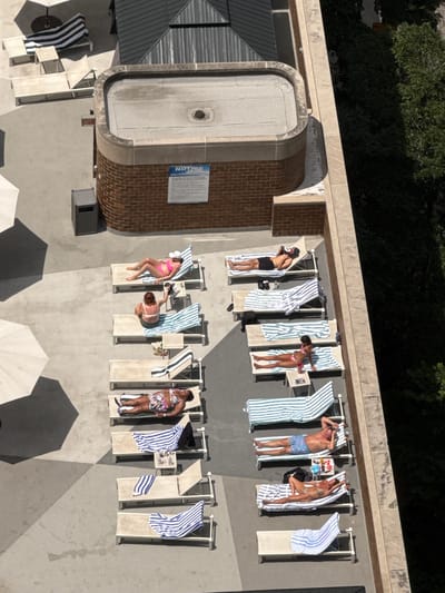 This is an aerial, high-angle, full shot of a rooftop deck in Chicago, Illinois, USA, on a sunny day. The deck is lined with lounge chairs, many of which are occupied by people sunbathing. Striped towels are draped over several chairs. In the foreground, a few people are visible, and further back, more lounge chairs are set up. Some of the chairs are empty. The background features trees and a brick building with a flat, grey roof. There is also a sign that says "NOTICE" attached to the brick building.