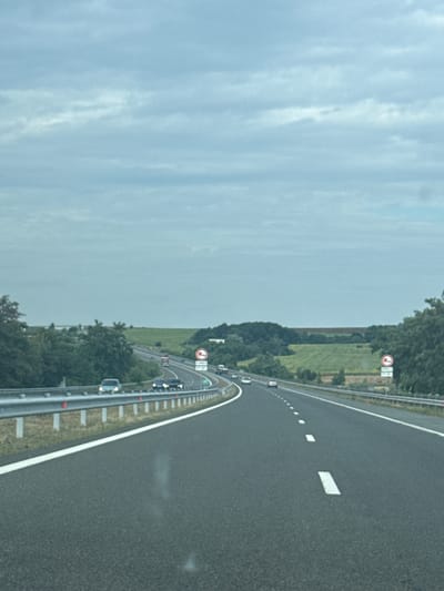 The image depicts a highway scene with a cloudy sky overhead. The road is asphalt with white lane markings, and a metal guardrail runs along the left side. Several cars are visible on the highway, traveling in the same direction. The landscape flanking the highway consists of green fields and trees. There are traffic signs on the right side of the road, including a circular sign with a red border and a diagonal red bar, commonly indicating a prohibition. The overall impression is that of a typical highway environment. There are no people or specific landmarks that would indicate the location is Zheleznik, Bulgaria.
