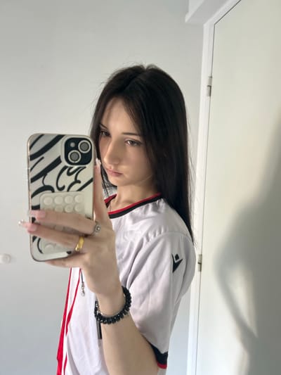 A young female is depicted taking a mirror selfie indoors in Cachan, France. She has long, straight dark hair with a center part and is positioned facing slightly right, looking towards the reflected smartphone screen. She wears a light-colored short-sleeved top with red and black trim around the neckline, featuring a black Macron logo on the right sleeve. Her left hand, adorned with a gold-colored ring on the ring finger and a silver-colored ring on the middle finger, holds a smartphone. The phone case is white with black, wavy, abstract patterns and a white, rectangular pop-socket featuring multiple circular indentations. Long, light-colored manicured fingernails are visible. On her right wrist, a black coiled bracelet and a thin metallic bracelet are worn. The background consists of a plain white wall or surface, with a white door and a visible door hinge on the right side of the frame.