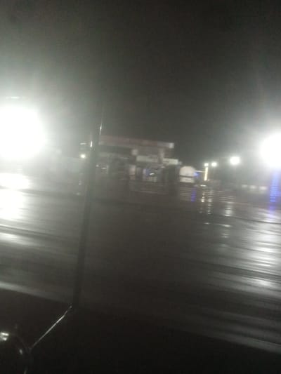 The image depicts a dimly lit, possibly rainy night scene at a gas station in Tundun Wada South, Nigeria. Bright lights from the gas station illuminate the wet pavement, creating reflections and a hazy atmosphere. The overall impression is one of low visibility and a nocturnal urban setting.