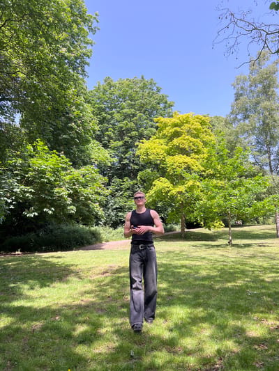 The image shows a person standing on a grassy area in Ipswich, United Kingdom. The person is wearing a black tank top, black pants, and sunglasses. They have their arms crossed. Behind the person are trees and a blue sky. The grassy area appears to be a park or garden.