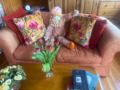 The image shows a living room in Golspie, United Kingdom. A light brown sofa is adorned with floral cushions in red and yellow hues, and two solid red pillows. A floral-patterned clown doll is seated on the sofa between the cushions. In front of the sofa is a wooden coffee table with a glass vase of red tulips, a basket of artificial white and yellow flowers, and two books. A wooden dresser stands against the wall in the background, while another wooden cabinet is on the left. The floor is hardwood.