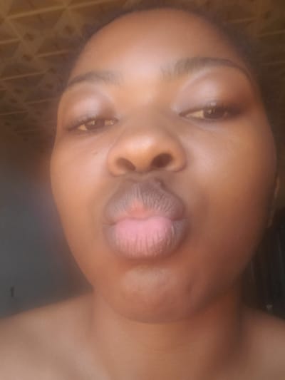 A close-up portrait features an individual, likely female, with a dark complexion, facing the camera directly. The subject has dark, textured hair visible around the top and sides of the head. Their dark eyes are open, with visible pupils and eyelashes. The eyebrows are well-defined. The subject's lips are significantly pursed into a "kissing" or "duck face" pose. No specific clothing or accessories are visible, only bare skin around the neck and shoulders.
The setting appears to be indoors. In the upper left background, a light-colored, patterned surface is visible, exhibiting a repeating geometric (possibly triangular or square grid) texture, which could be a ceiling or wall. The remainder of the background is indistinct, appearing darker and out of focus, suggesting a shadowed interior or plain dark surface. Lighting appears to be soft and frontal, illuminating the subject's face. The image does not contain any visual markers or details to confirm a specific geographical location such as Kakau, Nigeria.