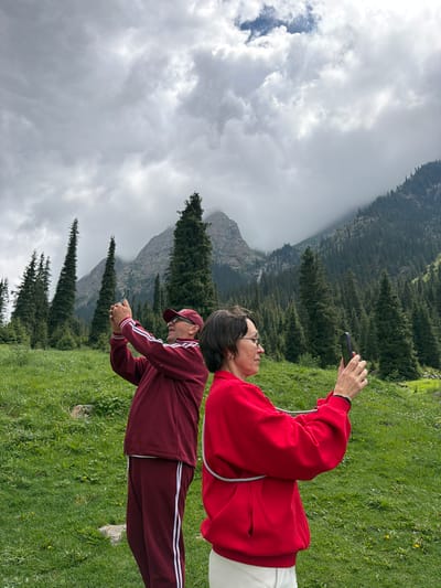 Two people are taking pictures of a mountain landscape with their phones. The person in the foreground, wearing a red sweatshirt and white pants, is holding a dark-colored phone horizontally. They are looking at the phone with a neutral expression and are positioned on a grassy hillside. Behind them, another person, wearing a maroon tracksuit and a baseball cap, is also looking at their phone, which they are holding in a similar fashion.
The background of the image shows a steep mountain slope covered with dense evergreen trees. Further in the distance, large, rocky mountains are visible, partially obscured by clouds. The sky is overcast with heavy gray clouds, suggesting a cool or possibly rainy day. The grass in the foreground is green and appears to be a meadow with a few small yellow flowers. The overall scene evokes a sense of exploration and appreciation for nature. No visible text is present in the image.