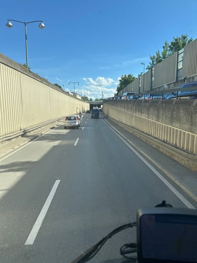 The image depicts a view from inside a vehicle traveling on a multi-lane road, likely an expressway or a highway, within Augsburg, Germany. The road is bordered by tall, ribbed concrete walls on both sides, which serve as sound barriers or retaining walls. The left wall is lighter in color and has a distinct ribbed pattern. The right wall is darker and also ribbed, with some windows or openings visible towards the top. Overhead lighting fixtures are present along the top of both walls.

The road itself is asphalt, with clear white lane markings. In the foreground, the dashboard of the vehicle is visible, with a small electronic device, possibly a GPS or dashcam, mounted on it. The road appears to be experiencing moderate traffic, with several cars and a van visible. A silver car with license plate "AIC-LW 315" is in the lane closest to the viewer on the left. Further ahead, a black van is visible in the right lane, and other vehicles are seen in the distance.

The sky is a clear, bright blue with a few scattered white clouds, suggesting fair weather. Trees are visible above the right-hand wall, indicating the presence of greenery in the surrounding area. The overall impression is of a typical urban or suburban road environment, potentially a section of an elevated road or a tunnel approach. The presence of specific car models and license plate formats could potentially aid in pinpointing the exact location within Augsburg, but without further context, it is difficult to be more precise.