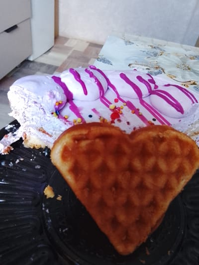 A heart-shaped waffle sits in the foreground on a black plastic plate, partially obscuring a slice of cake in the background. The cake is decorated with light purple frosting and a series of white dollops with purple stripes. Multicolored sprinkles are scattered on the cake. A white dresser is visible in the upper left corner, and a patterned tablecloth is behind the plate. The scene appears to be indoors. This is likely a residence in Kuyeda, Russia.