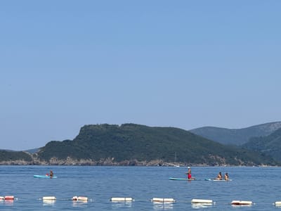In Pržno, Montenegro, the clear blue sea is dotted with a few people on stand-up paddleboards. In the foreground, a line of white buoys with red tops marks a section of the water. Further out, a person is paddleboarding on a blue board with a red cooler. To the right, another person in red shorts and a white hat is paddleboarding on a teal board, facing towards two other people on a paddleboard, one of whom is holding a fishing rod. In the background, a large, green, tree-covered hill rises from the sea, with smaller hills behind it, all under a clear blue sky. A sailboat is visible in the distance.