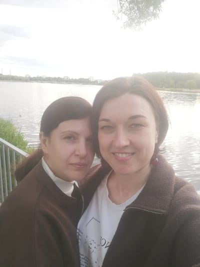 Two women are taking a selfie by a lake. The woman on the left has dark brown hair pulled back and is wearing a white turtleneck and a brown jacket. She is looking towards the camera with a neutral expression. The woman on the right has shorter brown hair and is wearing a white t-shirt with some text on it and a brown jacket. She is smiling at the camera. Both women appear to be in their 30s or 40s.
The background shows a body of water, with some trees and buildings visible on the opposite shore. There is also a railing in the foreground, suggesting the women are on a walkway or bridge. The sky is bright, with some clouds, indicating it might be daytime, possibly late afternoon, given the diffused sunlight. The overall scene is casual and relaxed. There is some text visible on the white t-shirt of the woman on the right, but it is not entirely legible. The location context is Kolpino, Russia.