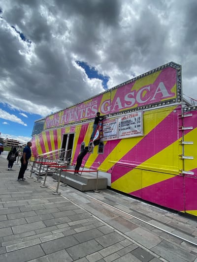 The image shows a view of a "Hnos Fuentes Gasca" carnival attraction, recognizable by its bright pink and yellow striped design. The carnival structure displays ticket information and show times in Spanish. A queue of people is waiting outside the attraction, separated by metal barriers with red ropes. Several people are visible: some are standing in line, while others are walking past. The ground is paved with gray bricks. The sky is cloudy, with patches of blue visible.