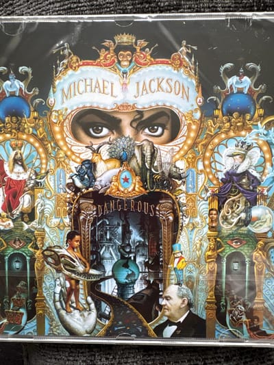 The image shows the cover art for Michael Jackson's album "Dangerous," rotated 90 degrees clockwise. The artwork is complex and fantastical, featuring Michael Jackson's face integrated into a collage of various symbols, figures, and scenes. The words "MICHAEL JACKSON" and "DANGEROUS" are visible within the design. The artwork is predominantly gold, black, and blue. The image does not show any scene located in the city of Manchester, United Kingdom.