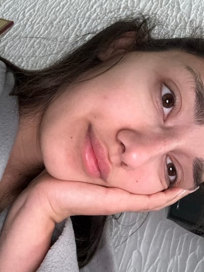 An individual, identified as an adult female, is depicted in a close-up, head-and-shoulders portrait, angled from a slightly overhead perspective. The subject is lying down or reclining, with her head tilted to the viewer's left. Her right hand supports her chin and lower jaw, with the palm making contact with the skin. Her gaze is directed towards the camera. She has dark brown hair, dark brown eyes with visible pupils and catchlights, and full lips that are slightly parted. Her complexion is natural, exhibiting minor moles or blemishes on the cheek. She is wearing a light gray, textured garment, visible on her left shoulder and arm. The background consists predominantly of a light-colored, quilted or textured fabric, consistent with bedding or upholstery. Partially visible in the top left corner is a dark object with a gold-colored edge, possibly a book. In the bottom right corner, a dark, rectangular object is partially obscured by the subject's hand and arm. The lighting is soft and frontal. The image itself provides no specific visual indicators to determine the location in Zankovići, Montenegro.