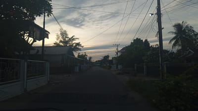 The image depicts a street scene in Meunasahteungkudigadong, Indonesia, likely during dusk or dawn due to the subdued lighting and cloudy sky. A paved road runs through the center, flanked by houses and vegetation. Utility poles with numerous wires crisscross the sky, and a street lamp is visible on one pole. On the left, a white metal fence with decorative patterns separates the road from a property with a building and trees. On the right, a similar fence encloses a property with dense foliage and a house. The overall impression is that of a quiet residential street in a tropical or subtropical climate.