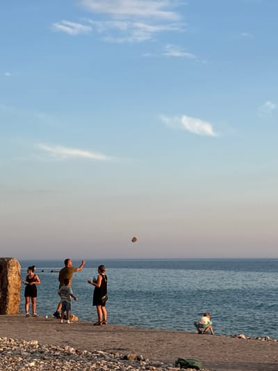 The image depicts a scene at the city of Perazića Do, Montenegro, with a focus on a group of people on a concrete embankment by the sea. In the background, the calm, blue sea stretches to the horizon, meeting a gradient sky with subtle clouds. The embankment is covered with pebbles and larger stones.
On the embankment, there are at least five individuals. On the left, a woman in a dark tank top and shorts stands near a large, rough-hewn stone. To her right, a man in a green t-shirt and shorts is captured mid-action, seemingly throwing or pointing towards an object in the air. Beside him, a young boy in a t-shirt and shorts watches. Further to the right, a woman in a black dress stands. In the distance, on the right side of the frame, a child is crouched down near the edge of the embankment, facing the water.
The sky above is a clear, pale blue, transitioning to a softer, warmer hue near the horizon, suggesting it is either sunrise or sunset. The overall atmosphere is relaxed and casual, with the people engaged in outdoor activity by the water.