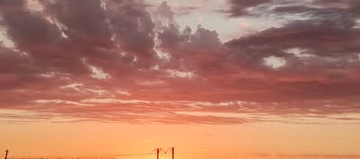 This media file captures a dramatic sky during what appears to be a sunset or sunrise. The dominant subjects are the clouds, which are a mix of deep purples and reds, transitioning into softer pinks and oranges as they descend towards the horizon. Some clouds are thick and textured, while others are thin and wispy, creating a dynamic visual effect.
The lower portion of the image shows a gradient of warm colors, ranging from a pale yellow near the horizon to a rich orange higher up. In the distance, there are hints of man-made structures, possibly power lines or pylons, silhouetted against the vibrant sky. One such structure, resembling an "M" shape, is visible in the lower right quadrant. The overall impression is one of natural beauty and perhaps a touch of industrial presence.
The time of day is clearly indicated by the warm, diffused light and the colors of the sky, pointing to either dawn or dusk. The weather appears to be clear enough to allow for a spectacular display of atmospheric color, though the presence of many clouds suggests some atmospheric activity. There are no people or animals visible in the frame. The setting appears to be an open landscape, given the vastness of the sky and the distant structures. The emotions evoked are likely awe, serenity, and perhaps a sense of wonder at the natural spectacle. There is no visible text in the image.