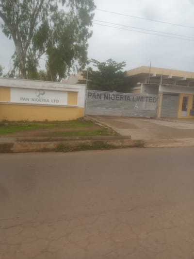 The image shows the front entrance of a facility labeled "PAN NIGERIA LIMITED" in Kakuri Gwari, Nigeria. The entrance features a large metal gate with horizontal slats. To the left of the gate is a white wall with a yellow section beneath it, displaying the "PAN NIGERIA LTD" logo and text. Behind the fence, a building with a pale yellow roof and white pillars is visible. Tall trees with green foliage are present on the left side of the image, in front of the building. The ground in front of the facility is a dirt pathway with some sparse grass. A paved road occupies the foreground of the image. The sky is overcast.