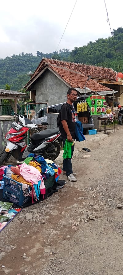 The image shows a man standing in front of a small shop in Kutawaringin, Indonesia. He is wearing a black t-shirt and blue jeans. He is holding a green bag in his right hand and a blue scarf in his left hand. He is looking at the camera. The shop has a sign in Indonesian that says "Fried Banana". There is a motorcycle parked in front of the shop. The motorcycle is red and white. There is a pile of clothing and a blue basket on the ground in front of the motorcycle. There is a pile of bananas on the table. There are green hills in the background. The scene is located at -6.974 degrees latitude and 107.519 degrees longitude.