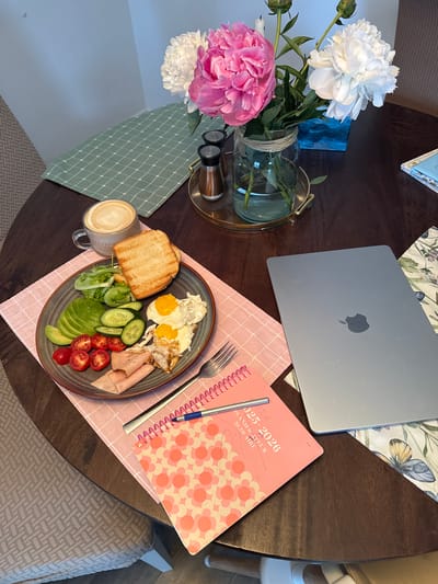 A overhead shot shows a round, dark brown table set for a meal and work. In the foreground, a pink, floral-patterned notebook with a blue pen lies open. Next to it is a closed, silver laptop computer with an Apple logo. To the left of the laptop, a plate holds a breakfast meal consisting of sliced avocado, cherry tomatoes, cucumber slices, lettuce, ham, and two fried eggs, accompanied by two pieces of toasted bread and a cup of coffee with latte art. Behind the plate, a glass jar filled with water holds a bouquet of pink and white peonies. A green, checkered placemat and a gold tray with salt and pepper shakers are visible behind the flowers. The table is set on a patterned rug, and a chair with a woven texture is partially visible on the left side of the image. The scene is well-lit, suggesting an indoor setting. The specific location in Gatineau, Canada cannot be determined from the image.
