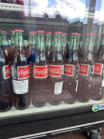 The image shows a refrigerated display case filled with glass Coca-Cola bottles, each labeled with "NO RETORNABLE" (non-returnable) and "HECHO EN MEXICO" (made in Mexico). The bottles have red and white labels with the Coca-Cola logo and nutritional information. The bottles are stacked horizontally in multiple rows. The display case has a black frame and a clear glass front, reflecting the sky and nearby buildings. The location is Lake Buena Vista, United States, determined by the Innovatus logo on the cooler, indicating its origin.