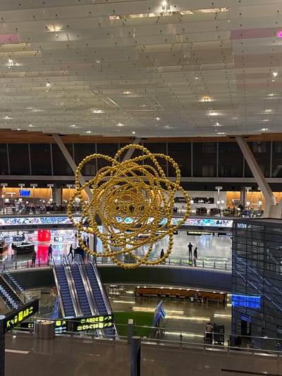 The image shows a view of the interior of Hamad International Airport in Doha, Qatar. A large, intricate golden sculpture hangs from the ceiling, composed of interconnected circular strands made of small golden spheres. Below the sculpture, the airport concourse is visible, with escalators, walkways, and seating areas. Several people are walking and standing on different levels of the concourse. Shops and restaurants are situated on a lower level, identifiable by signage such as "THE JUICE." The ceiling is a modern, expansive structure with many rectangular panels and lights. The overall environment is bright, spacious, and modern.