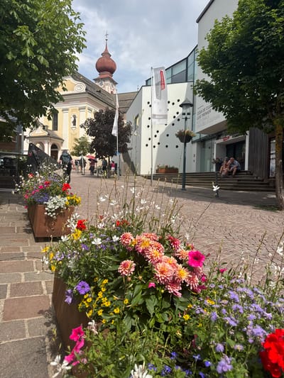 The image depicts a street scene in Ortisei, Val Gardena, Italy. In the foreground, large planters overflow with a vibrant assortment of flowers, including pink and orange dahlias, purple petunias, yellow daisies, and delicate white blossoms. The planters are made of a dark, rustic metal. The ground is paved with cobblestones.
In the mid-ground, a street extends into the distance, lined with buildings. On the left, a yellow and pink church with a red onion dome and clock tower is visible, partially obscured by trees. Adjacent to the church is a modern white building with a glass facade and a prominent flag bearing the text "Dolomites Val Gardena". Another building to the right is also modern, with a large glass entrance and stairs leading up to it. On these stairs, two individuals, a man and a woman, are seated facing each other.
In the background, several people are walking along the street. One person is riding a bicycle. A red umbrella is held by someone in the crowd. The sky is partly cloudy. The overall impression is of a pleasant, well-maintained town square on a sunny day.