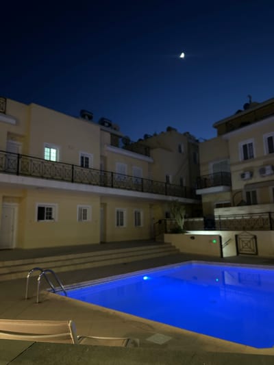 This image, taken at night in Peyia, Cyprus, depicts a well-lit swimming pool area adjacent to a multi-story building. The pool, illuminated by a vibrant blue light from underwater fixtures, reflects the surrounding architecture and the dark, clear night sky. A crescent moon is visible in the upper portion of the frame. The building, painted a light yellow, features multiple windows and balconies with black metal railings. Some windows are illuminated from within, suggesting occupancy. Two lounge chairs are partially visible in the foreground, placed on a paved surface. A metal pool ladder with two handles is positioned at the edge of the water. The overall scene conveys a sense of tranquility and resort-like atmosphere.