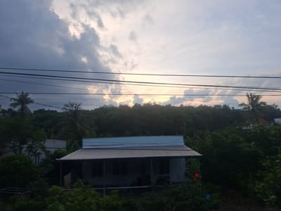 This image captures a serene scene in Dong Nai City, Vietnam, likely during the late afternoon or early evening. The sky is filled with dramatic, textured clouds, with patches of bright sunlight breaking through, suggesting a dynamic weather pattern. The lighting indicates either sunrise or sunset, casting a warm glow on the clouds and the landscape.
The foreground is dominated by power lines stretching horizontally across the frame, creating a sense of depth and hinting at human infrastructure in this semi-rural or suburban setting. Below the power lines, a modest, single-story house with a light blue roof and a dark awning is visible. The house appears simple, with dark windows suggesting the interior is either not illuminated or the angle of the sun makes it appear so. Some colorful lights, possibly small LED decorations, are visible on the awning of the house.
Behind the house, a dense, dark green forest or tree line forms a backdrop. Palm trees are interspersed within the foliage, a characteristic feature of tropical or subtropical locations. The overall impression is one of quietude and natural beauty, with the man-made elements integrated into the landscape. The lush greenery and the tropical trees firmly place the scene in a warm climate. The weather appears partly cloudy, with the sun making an effort to shine through. There are no visible people, and the house appears unoccupied or in a state of repose. The mood is peaceful and slightly melancholic, due to the subdued lighting and the imposing presence of the clouds.
