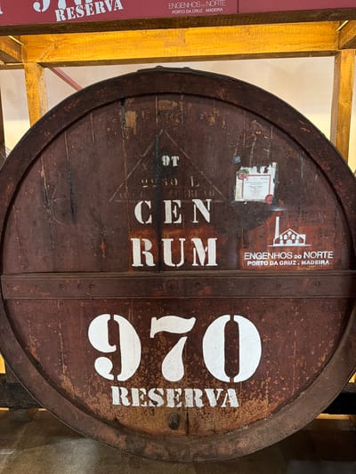 A medium shot depicts a large wooden barrel with prominent white lettering and markings. The words "CEN RUM" are stacked vertically in the center. Above them, a smaller logo with the text "ENGENHOS DO NORTE" and "PORTO DA CRUZ. MADEIRA" (the city of Porto da Cruz is within the Machico municipality of Portugal) can be seen along with what appears to be a small information label. To the right, the large number "970" is vertically aligned alongside the word "RESERVA". The barrel has metal bands and rivets holding it together, and the wood shows signs of aging and wear. The barrel is likely used for storing or aging rum. Part of what appears to be a wooden frame and the corner of a display with the word "RESERVA" is visible on the left side of the frame.