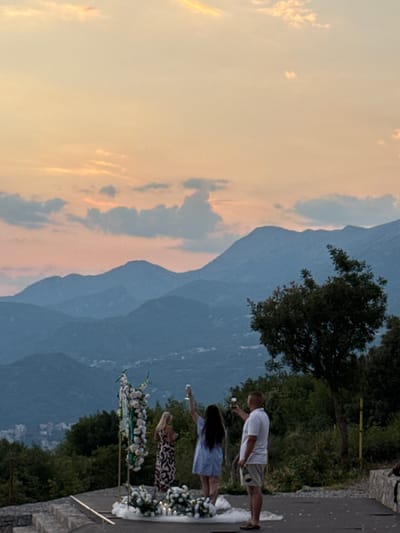 This media file captures a wedding ceremony held outdoors in Čelobrdo, Montenegro, at dusk. The sky is filled with soft, warm hues of orange and pink, indicating the sunset. In the foreground, a man in a white polo shirt and khaki shorts is visible, holding up his phone as if to record or take a photo. To his left, a woman with long dark hair wears a light blue dress and also appears to be holding a phone. Further to the left, another woman with blonde hair is dressed in a patterned dress and holds a phone.

Behind the people, a dramatic backdrop of lush green trees and imposing, layered mountains is depicted. The mountains fade into a hazy blue in the distance, suggesting a vast and picturesque landscape. A decorative floral arch, adorned with white flowers, stands near the women, suggesting it's part of the wedding setup. The ground appears to be a paved pathway or road, and a stone wall is visible on the right side of the frame. The overall atmosphere appears serene and celebratory, with the golden light of the sunset casting a warm glow over the scene. No visible text is present in the media file.