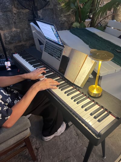 A person's hands are visible on the keys of a black digital piano. The person is seated on a grey stool and is wearing a dark blue shirt with a white floral pattern and black pants. A laptop displaying sheet music is propped up on a white stand behind the piano. To the right of the piano, a golden desk lamp with a round shade illuminates a music book. The scene appears to be outdoors, with a stone wall and lush green foliage in the background. A white wooden table with a dark green runner is visible to the right of the piano. The ground is paved with grey stones. The location is Dobrota, Montenegro.
