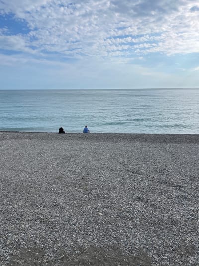 The photograph depicts a coastal scene in Olginka, Russia. The image is vertically oriented. A cloudy blue sky occupies the upper left corner. The sea stretches across the central portion of the frame. Two figures are present near the water's edge. One is wearing a light blue shirt and is standing. The other is wearing dark clothing and is crouching. A pebble beach fills the right side of the image.