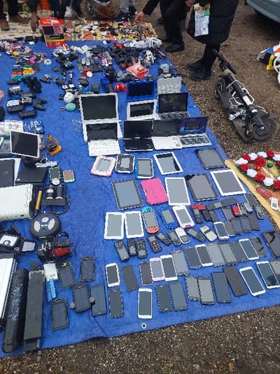 The image depicts an outdoor market stall in Eckbolsheim, France, showcasing a large array of used electronic devices meticulously arranged on a royal blue tarp laid over a gravel ground surface. The upper portion of the tarp features various laptops, both open and closed, alongside smaller electronics such as gaming controllers, smartwatches, and assorted chargers. The mid-section displays numerous tablets, some enclosed in colorful protective cases. The lower two-thirds of the tarp are densely covered with multiple horizontal rows of mobile phones, encompassing a wide range of smartphones in various models and colors, interspersed with older feature phones. Other visible objects include several black solar power banks, a portable air pump, and a white game console unit. In the upper right quadrant, a black electric scooter rests on its side adjacent to a small arrangement of artificial red and white roses. Several partially visible individuals are present in the background, standing near the stall, with one person's legs, lower torso, and arm distinctly visible near the scooter and the displayed items.