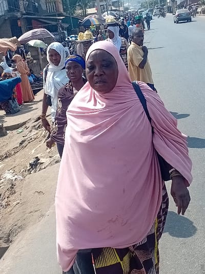 Several women wearing traditional Muslim clothing walk along a street. The foreground subject wears a pink hijab, while others are in white and yellow. They are on a paved surface, potentially a road, near a bustling area with buildings, stalls, and people in the background. The scene seems to capture a daily moment in a vibrant, busy neighborhood.