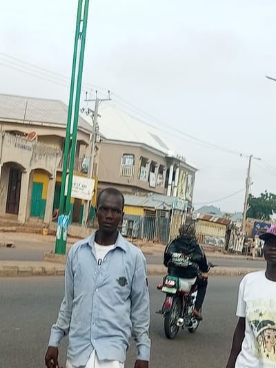The image depicts a street scene in Garko, Nigeria. In the foreground, a man wearing a light blue button-down shirt and white pants stands looking at the camera. To his right, another person wearing a white t-shirt with graphics and a purple hat is partially visible. In the midground, a person on a red motorcycle wearing a black jacket with white lettering is riding away from the camera. The background shows a street with several buildings, some with balconies and storefronts. There is also a green pole with wires and a utility pole with insulators. The sky is overcast.
