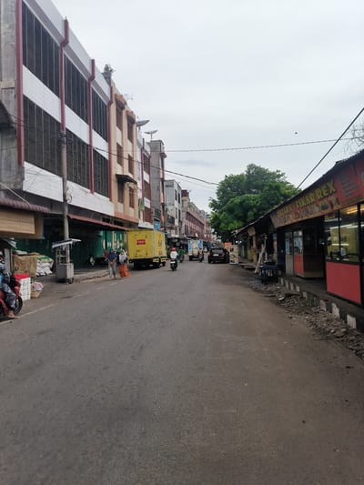 The image depicts a street scene in the city of Bandarbireuen, Indonesia. On the left, a multi-story building with white tiled facade and red accents lines the street. A bamboo-slatted awning extends from the building, with stacked white sacks and a basket visible beneath it. A person on a red motorcycle is parked in front of this structure. Further down the street, two women in headscarves are walking towards a yellow truck. Several shops with green shutters are visible along the sidewalk. On the right side of the street, a row of food stalls with red and yellow signage stretches into the distance. One stall, labeled "QATARY / FIKAR REX", displays menus and images of food. A dark-colored car is parked on the street. A motorcycle is moving away from the camera on the right side of the road, and another motorcycle is visible further down. A large green tree stands behind the food stalls. The sky is overcast, and power lines crisscross above the street. The road surface is asphalt, with scattered debris along the right curb.