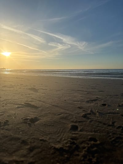 This is a low-angle, wide-shot photograph of a sandy beach at sunset, with the ocean stretching to the horizon. The sun, a bright orb, is setting on the left side of the frame, casting a warm, golden glow across the scene and creating a lens flare effect. Thin, wispy clouds streak across the sky, tinged with yellow and orange from the setting sun. The water is calm with gentle waves lapping the shore. The sand in the foreground is textured with footprints and small pebbles. In the distance, on the left, a pier or lighthouse structure is visible. The overall impression is one of tranquility and natural beauty. The image is representative of the beaches found in Ustka, Poland, a coastal town known for its picturesque shoreline and sunsets.