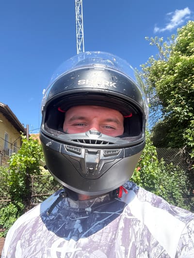 A close-up, front-facing selfie shows a person wearing a black and clear-visored motorcycle helmet. The helmet has a "SHARK" logo visible on the top. The person's eyes and forehead are visible through the visor. They are wearing a camouflage-patterned shirt. The background is outdoors, with a bright blue sky and lush green foliage, including trees with white flowers. To the left, a portion of a light-colored building with a terracotta roof is visible. A tall, white metal structure, possibly a radio tower or crane, is also visible in the upper part of the image. The scene appears to be set in a residential area within the city of Radomir, Bulgaria.