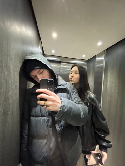 An image captured in Cachan, France, depicts two individuals reflected in a mirror, likely within an elevator. The environment features dark, vertically textured panels on the left, and horizontally brushed metallic panels in the background, topped by a reflective white ceiling with two recessed circular lights.
In the foreground, a male subject, dressed in a black hooded puffer jacket with the hood up over a black beanie, leans against the left wall. He holds a dark-colored smartphone in his right hand, positioned to capture the reflection. A gold ring is visible on his right ring finger.
Behind and to the right of the male is a female subject with long, dark hair. She wears a black jacket, possibly leather, and her lips are pursed in a pouting expression. She holds a lighter-colored smartphone in her right hand, which displays a reflection of the scene on its screen. The individuals are in close proximity, with the male actively taking the photograph.