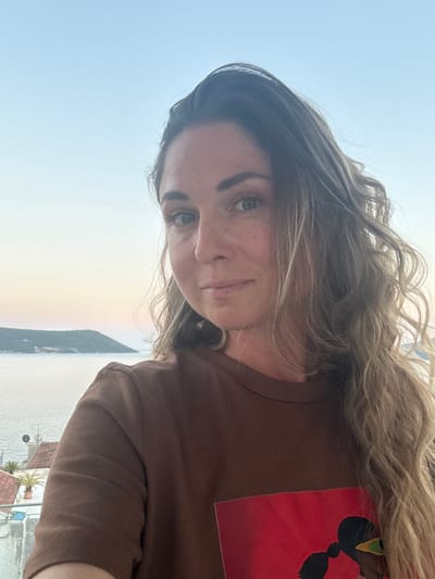 A woman with long, wavy blonde hair and green eyes is captured in a selfie, looking towards the camera. She is wearing a brown t-shirt with a red rectangular graphic on the lower portion, featuring a silhouette of a person with braided hair and a green and yellow detail. Her skin is lightly tanned and has freckles. The background suggests an outdoor setting in Herceg Novi, Montenegro, with a body of water and a green, hilly landmass in the distance. The sky is a pale blue, indicating it could be dawn or dusk. Part of a building with red-tiled roofs and white walls is visible to the left of the frame, with a palm tree and satellite dishes on its rooftop.