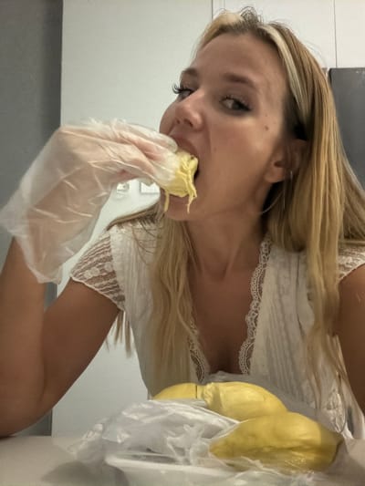 A young woman with blonde hair is eating durian. She is wearing a white lace top and plastic gloves. The durian is yellow and wrapped in plastic. The scene appears to be indoors, possibly in a kitchen or dining area. The lighting suggests daytime. The woman's expression is difficult to discern due to the angle of the photo, but she seems focused on eating the fruit. The background is out of focus, but appears to be a neutral colored wall. The overall impression is a candid shot of someone enjoying a tropical fruit.